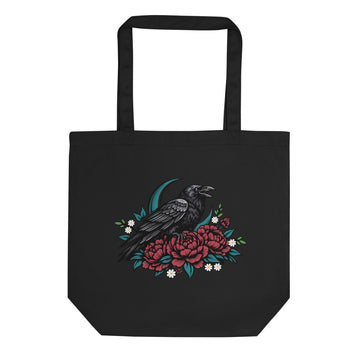 A black canvas tote bag with a graphic print of a black raven perched on red flowers with green leaves and small white blossoms, shown against a plain white background.