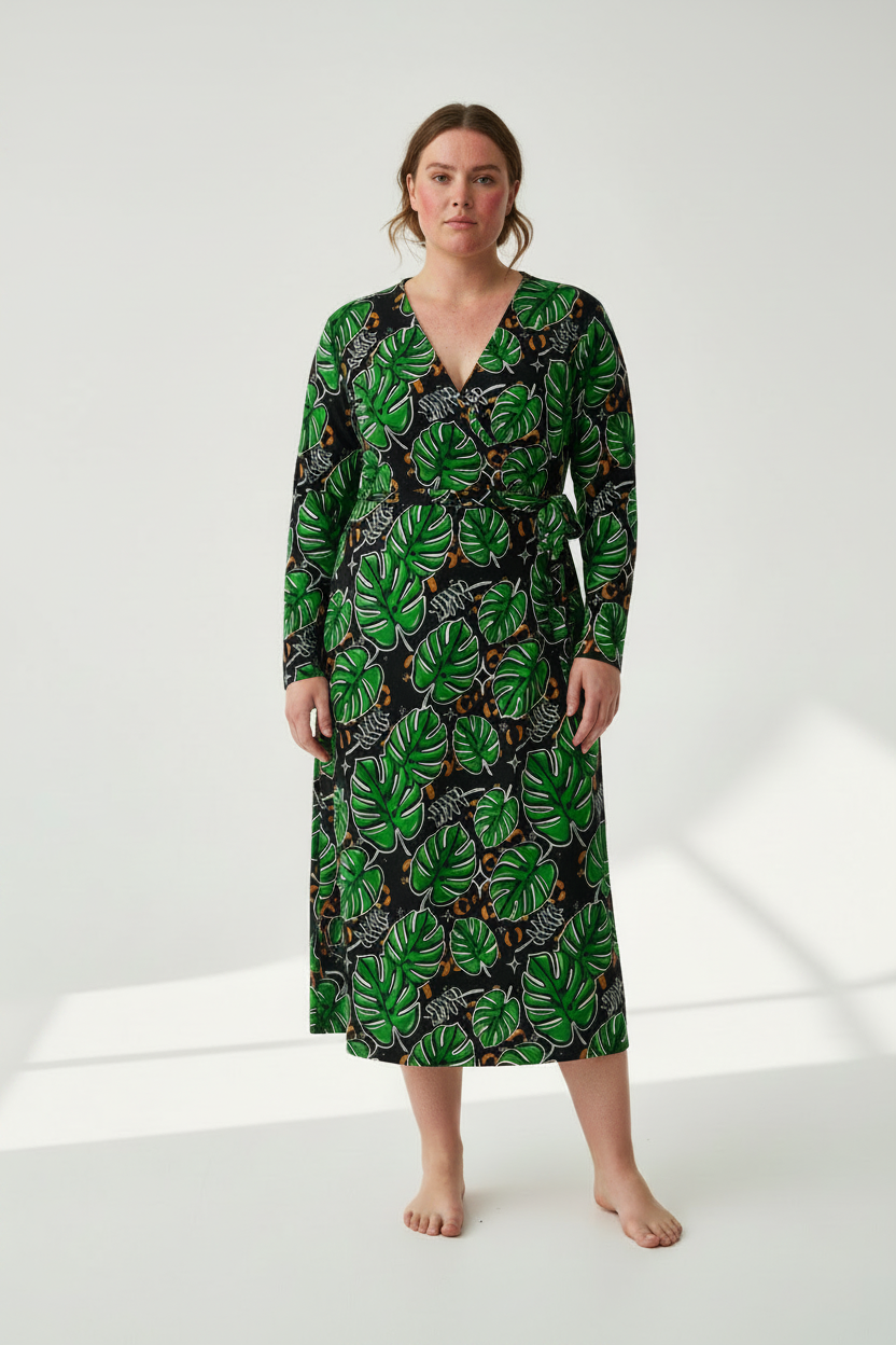 A woman modeling a long-sleeve midi wrap dress with a green tropical leaf print on a black background, standing against a white studio backdrop.