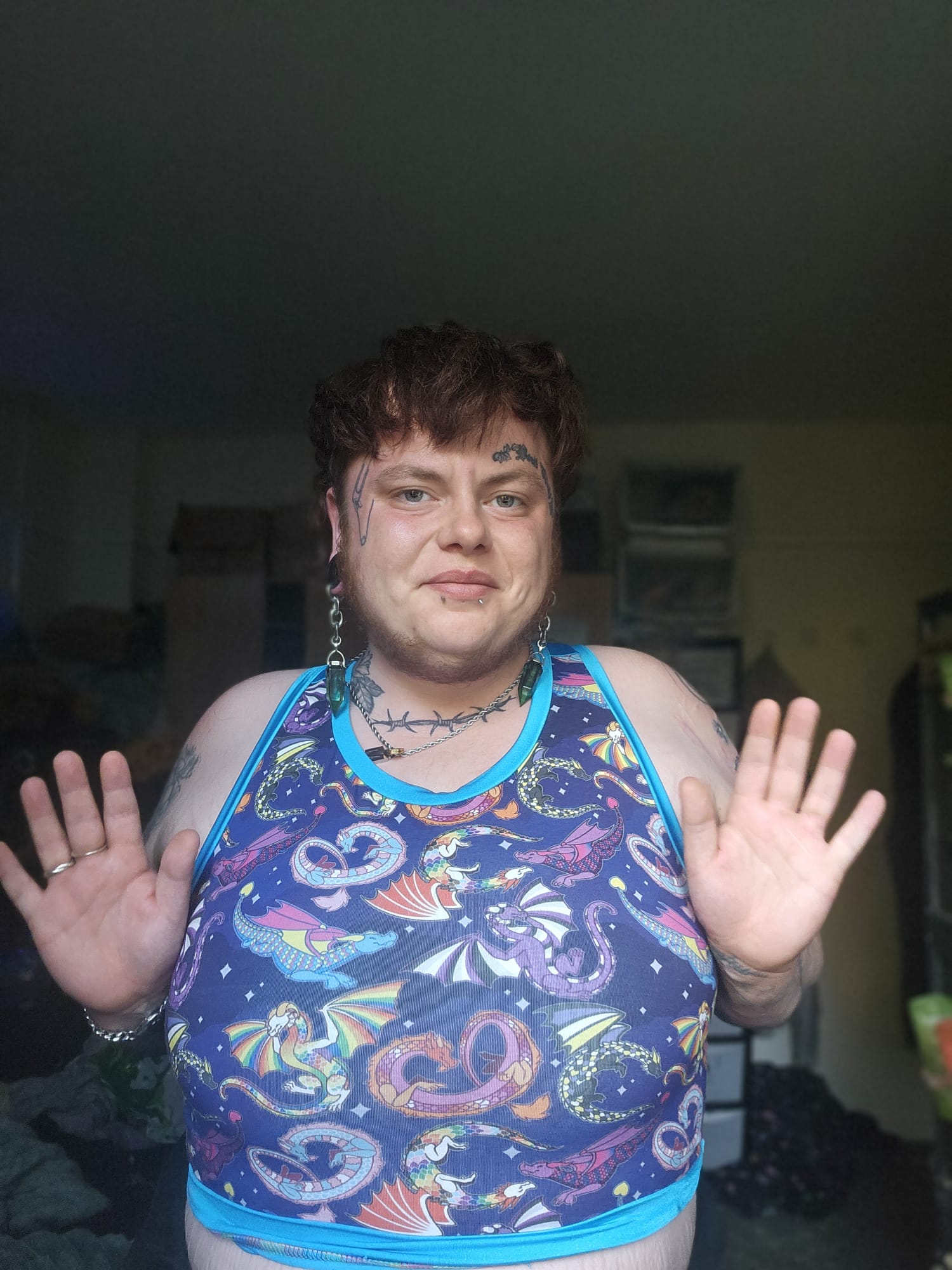 A person wearing a sleeveless binder tank top with a colorful mermaid print on a dark blue background, standing indoors with both hands raised.