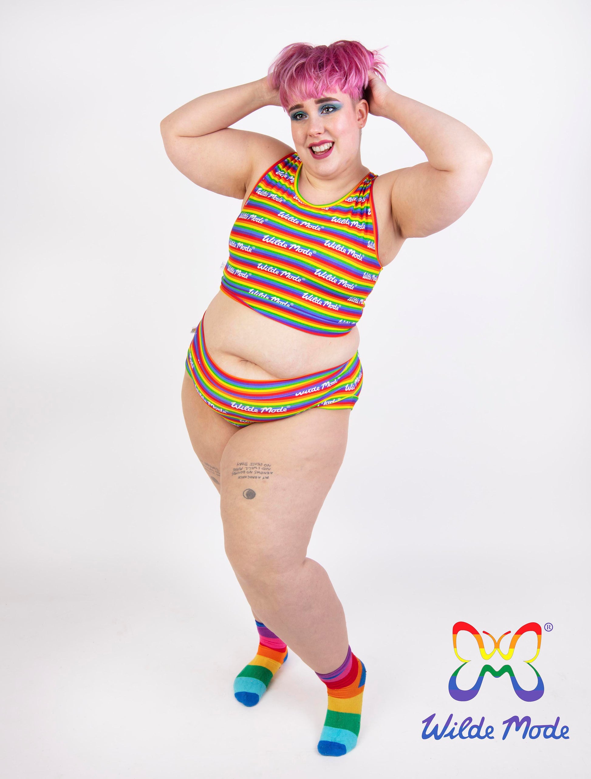 A plus-size model with pink hair poses in a rainbow-striped bikini top and matching bottoms, paired with rainbow-striped socks, on a white studio background. The 'Wilde Mode' logo is visible in the bottom right corner.