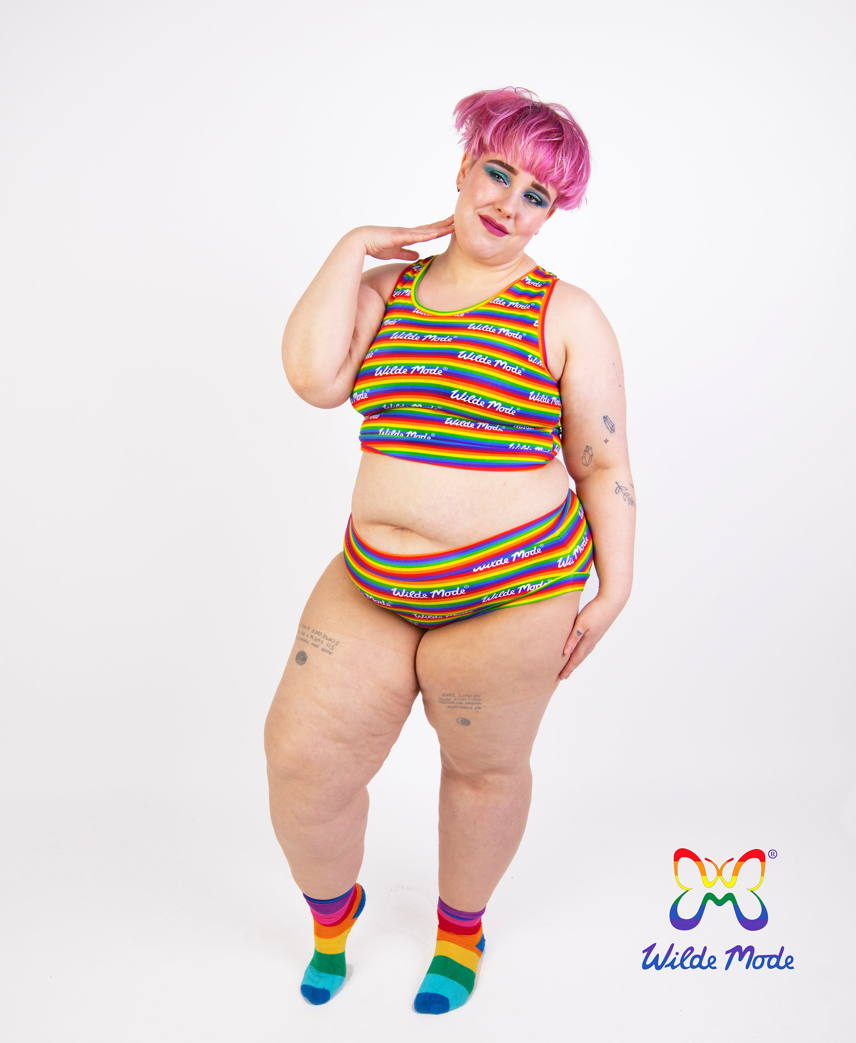 A plus-size model with pink hair wearing a rainbow-striped crop top and matching bikini bottoms, paired with matching rainbow socks, against a white studio background. The Wild Mode logo is visible in the bottom right corner.