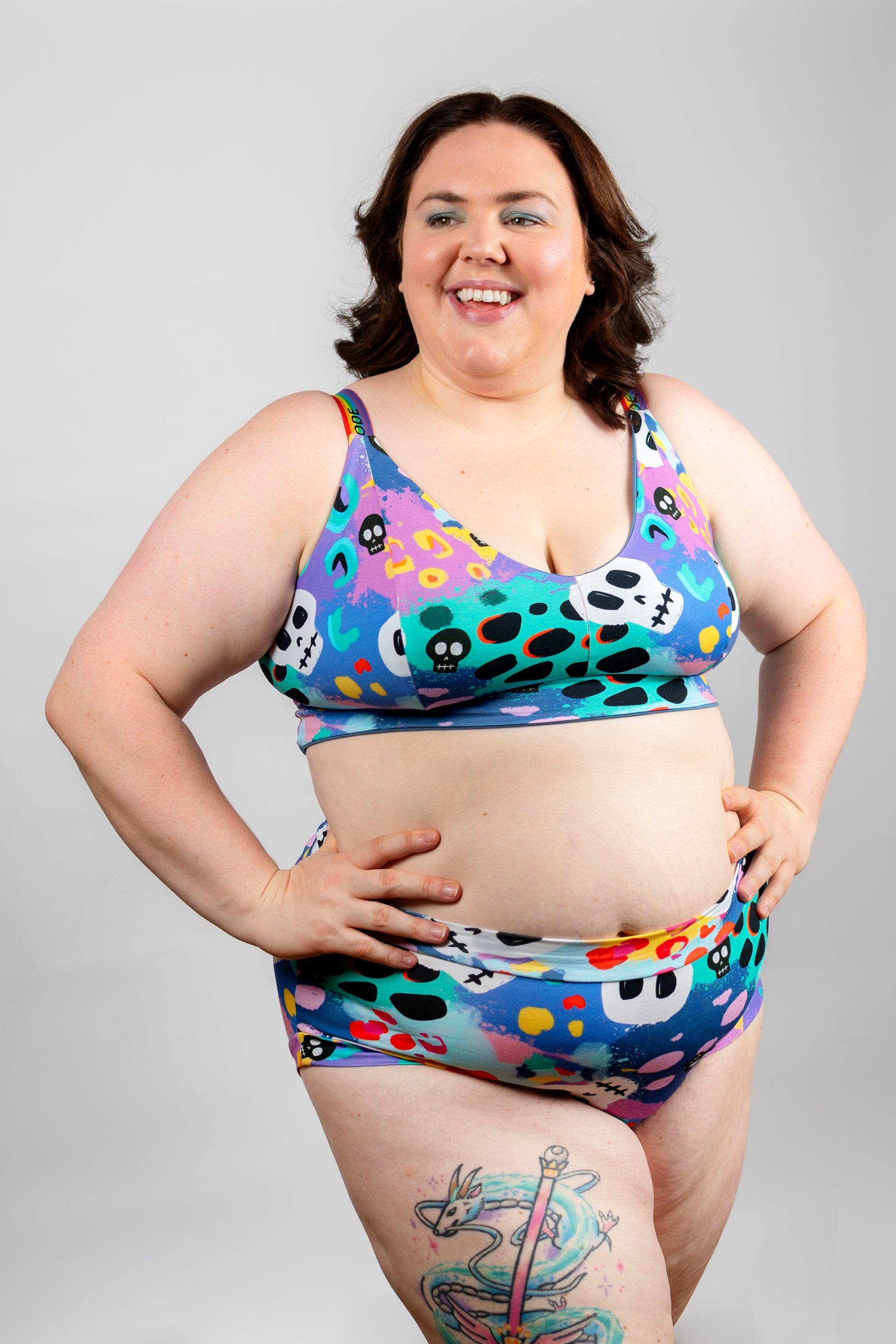 A woman modeling a two-piece bikini set with a colorful, playful pattern of skulls, paint splatters, and dots on a light gray studio background.