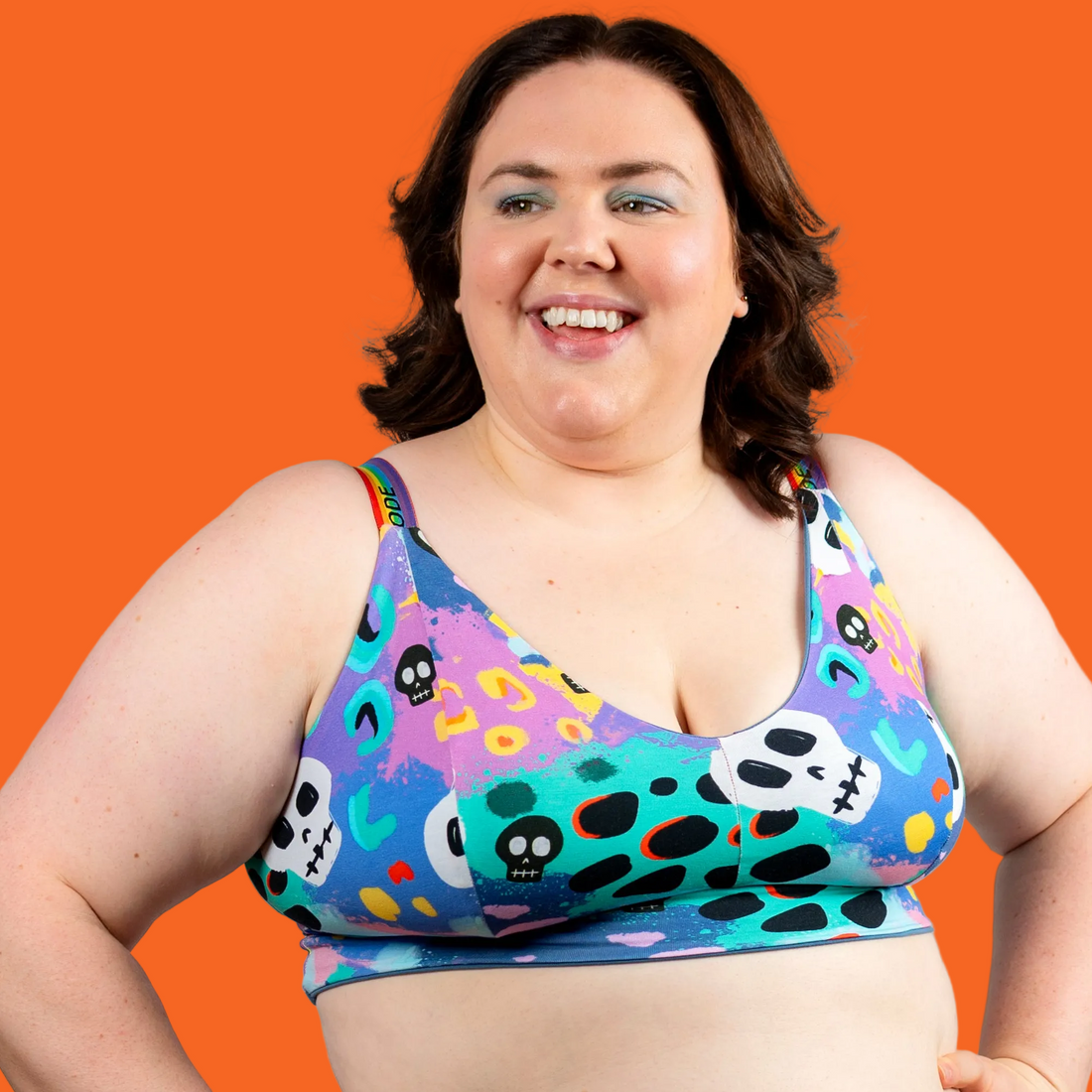 A smiling woman wearing a colorful patterned sports bra with a vibrant multicolor print of skulls, dots, and abstract shapes on a blue, purple, and pink background, set against a plain light gray studio backdrop.