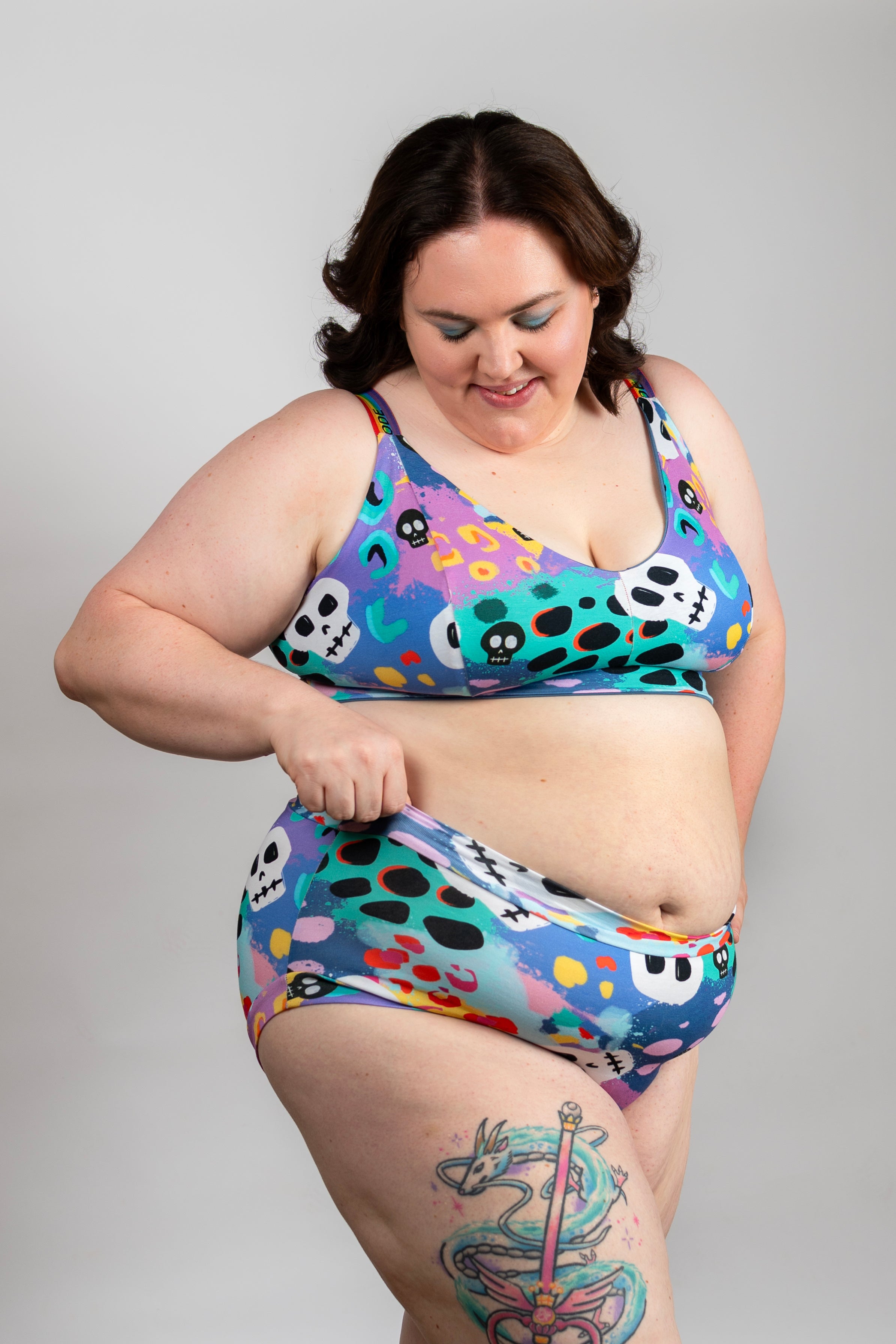 A plus-size woman modeling a matching two-piece bikini set with a colorful, playful pattern of skulls, eyes, and dots on a purple, blue, and teal base. She is standing against a solid light gray background.