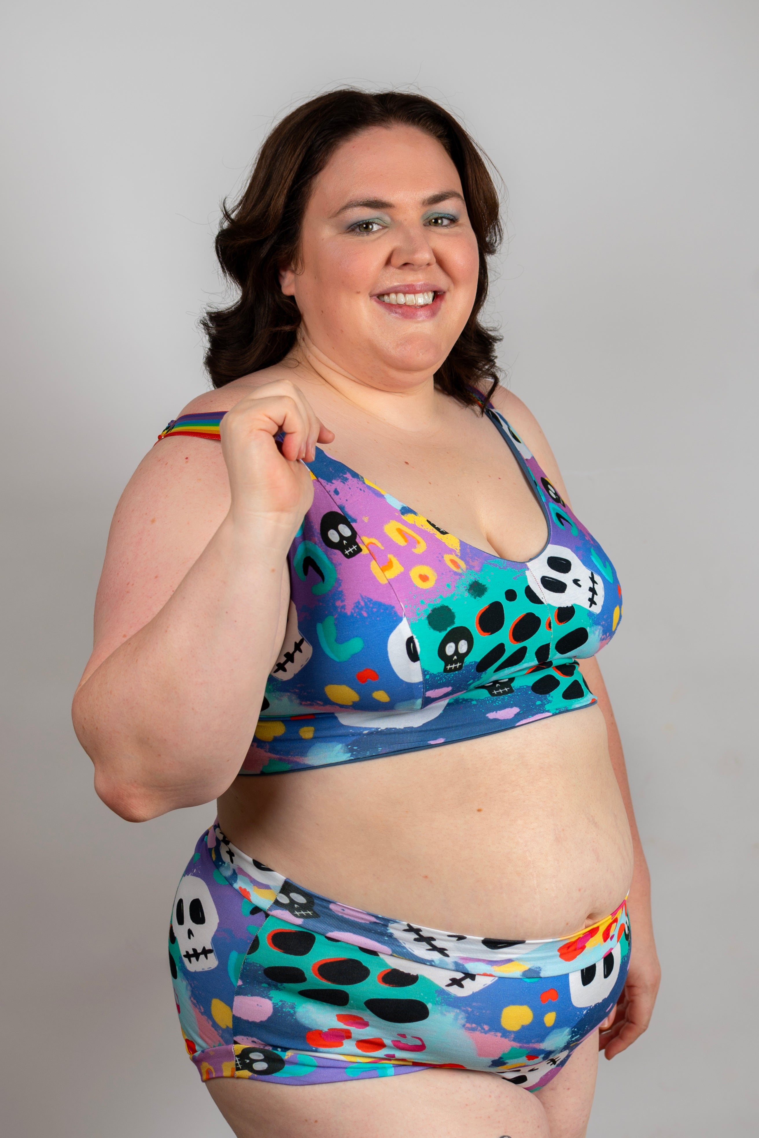 A woman modeling a two-piece bikini set featuring a colorful, playful pattern of skulls, dots, and abstract shapes in blue, purple, green, yellow, and black, set against a plain light gray background.