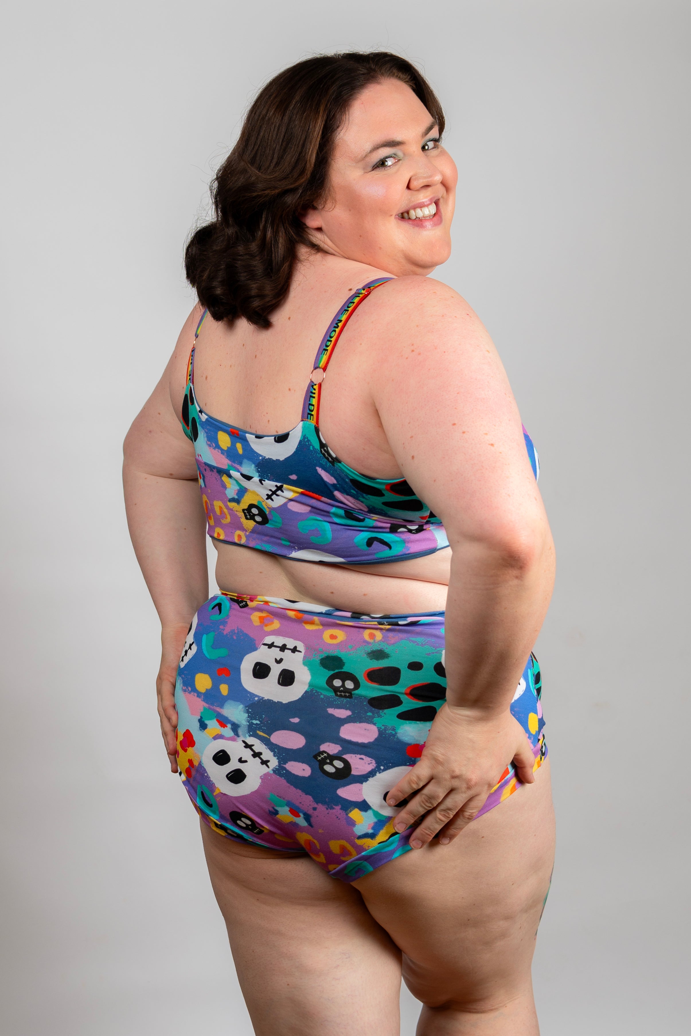 A woman modeling a two-piece swimsuit with a colorful sugar skull pattern, photographed against a neutral gray studio background.