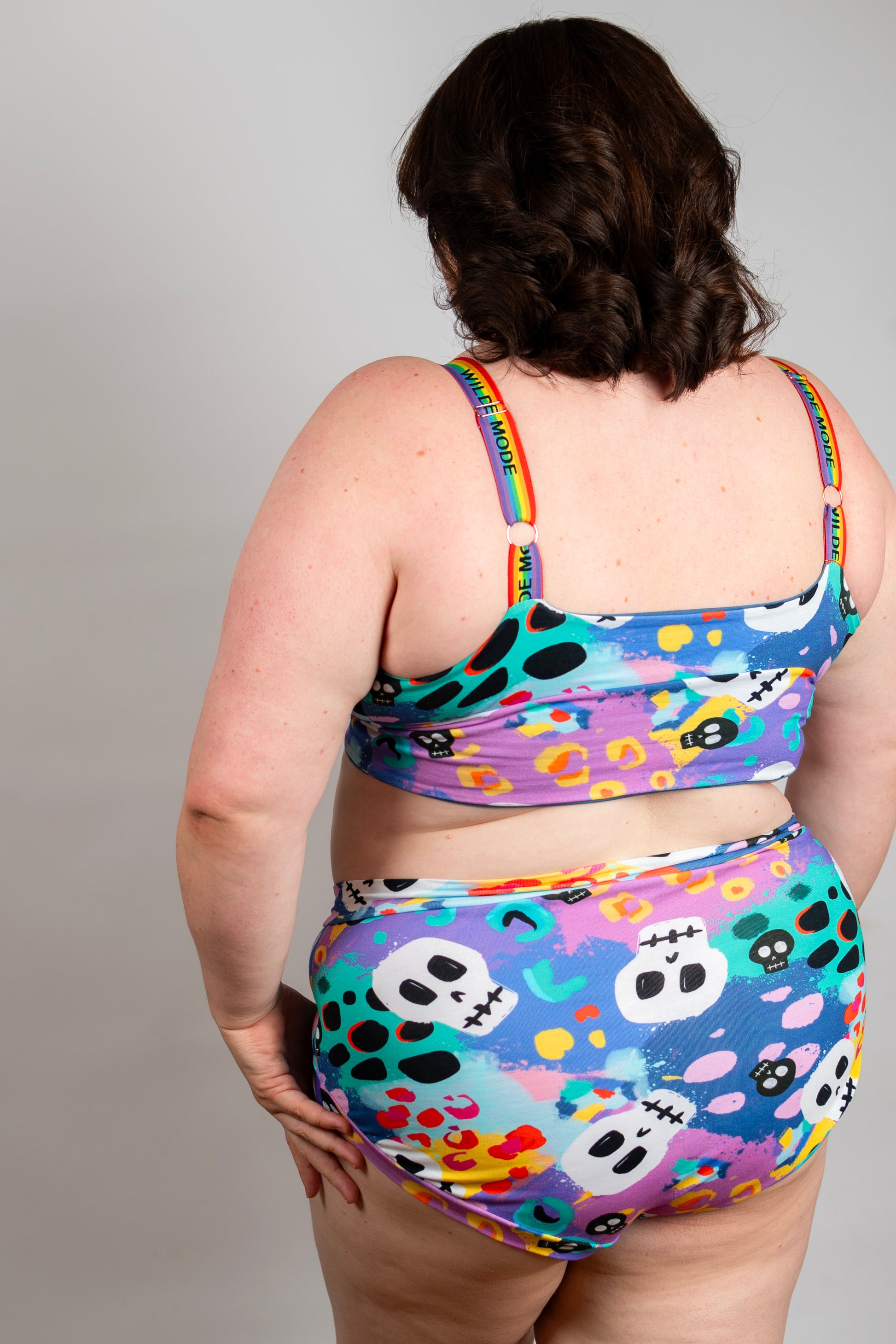 A woman shown from behind wearing a colorful two-piece swimsuit with a Day of the Dead style print featuring skulls, ghosts, and floral motifs on a purple, blue, and teal background.