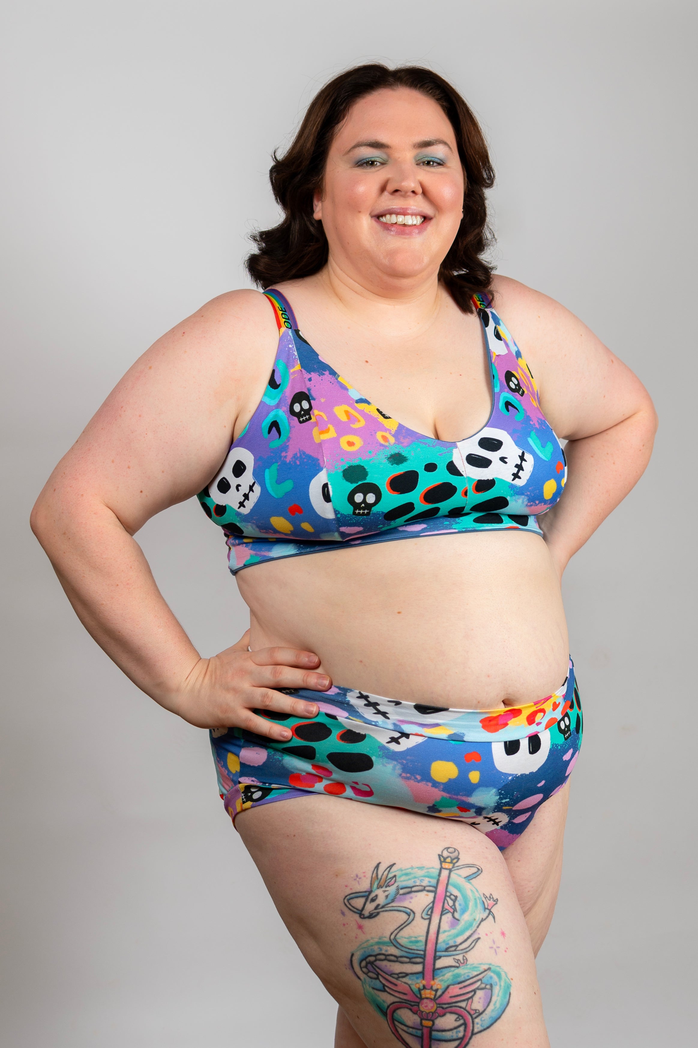 A woman modeling a two-piece bikini swimsuit with a colorful, busy pattern of skulls, hearts, and dots in shades of blue, purple, pink, yellow, and black, set against a plain light gray background.