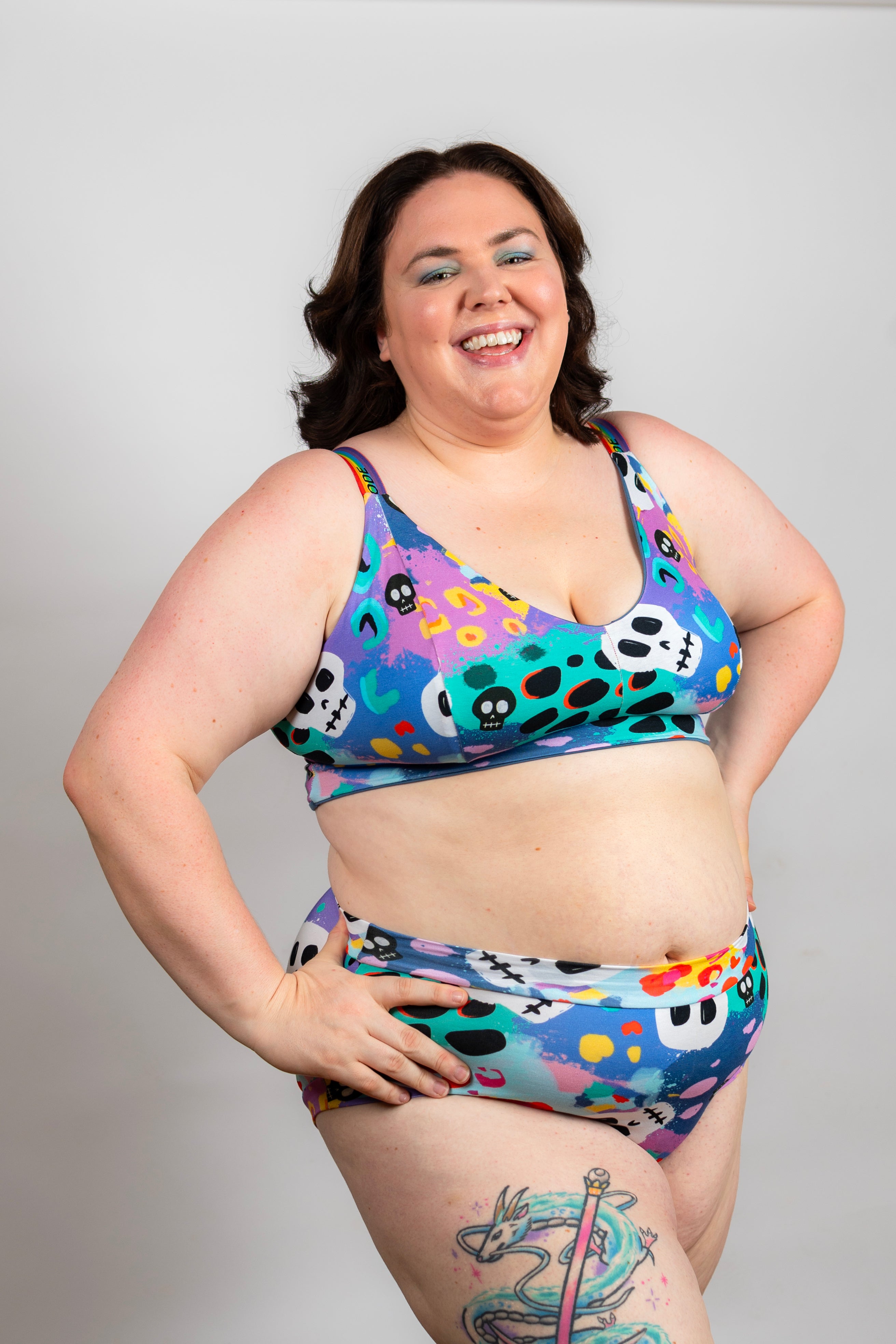 A woman modeling a two-piece bikini set with a colorful, playful pattern of skulls, hearts, and abstract shapes on a light gray studio background.