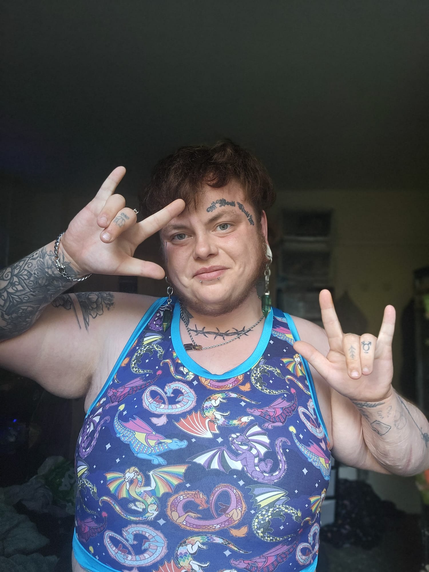 A person wearing a blue binder top with a colorful psychedelic print of dragons, hearts, and swirls, posing with both hands raised in the rock on. The background is a dark, cluttered indoor space.